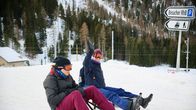 Kaksi ihmistä hiihtolasit päällä kelkkojen päällä lähdössä laskemaan mäkeä. Taustalla näkyy alppirinteen kuusimetsää ja juna-asema, valkomustat kyltit osoittavat parkkipaikalle ja junalle.