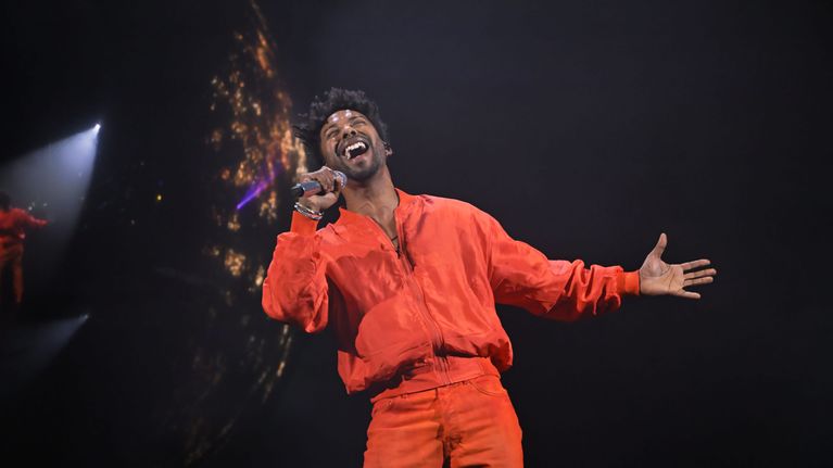 John Lundvik i orange jbyxor och orange jacka, sjunger i mikrofon mot svart bakgrund.