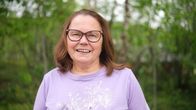 Henkilö seisoo ulkona vihreän kasvillisuuden keskellä, päällä vaalea violetti paita, jossa valkoinen kukkakuosi.