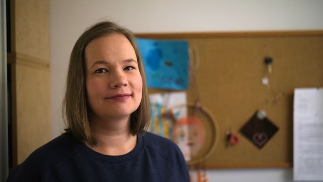 Sivukuva ihmisestä. Hän hymyilee vienosti. Taustalla näkyy ilmoitustaulu.