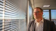 Silmälasipäinen, valkoiseen kauluspaitaan  ja tummaan puvuntakkiin pukeutunut mies katsoo korkean rakennuksen ikkunasta ulos, aurinko osuu hänen kasvoilleen. Ikkunasta näkyy kaupunkimaisemaa ylhäältä päin.