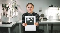 Emilia Laatikainen katsoo suoraan kameraan ja pitää käsissään piirtämäänsä kuvaa itsestään.