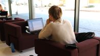 En person sitter i en fåtölj och arbetar vid en dator intill ett stort fönster.