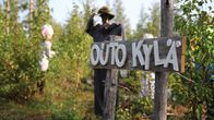 Metsän keskellä on itsetehdyn näköinen puinen kyltti, jossa lukee outo kylä. Taustalla metsässä epätarkkoja ihmishahmoja.