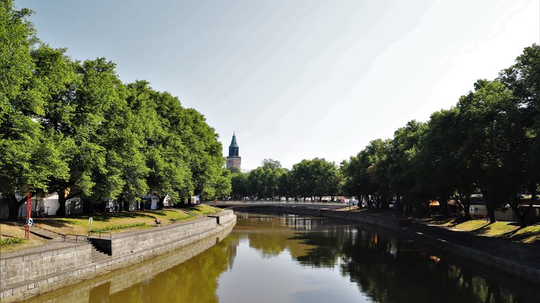 Aura Å mitt i sommaren. På bilden syns grönskande träd, biblioteksbron och domkyrkan i bakgrunden. Fotot är taget från Aurabron. 