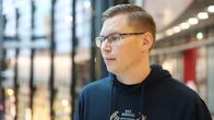 Mies seisoo sisätiloissa, päällään tummansininen huppari, jossa on painatus. Taustalla on lasiseinä ja kauppakeskus.