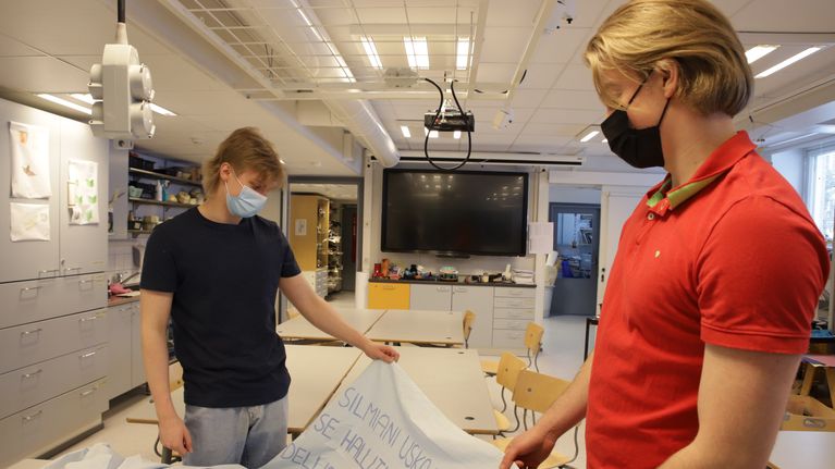 Abiturientit Santeri Lehtiniemi ja Veikka Ruotsalainen katsovat kuvaamataidon luokan pöydälle levitettyä abikuorma-auton kylkeen kiinnitettävää lakanaa. Abeilla on maskit kasvoillaan.
