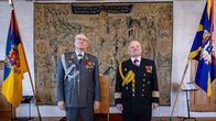 Två uniformerade män med medaljer står framför en vävd tapet, flankerade av flaggor i en högtidlig sal med trägolv och träpaneler.