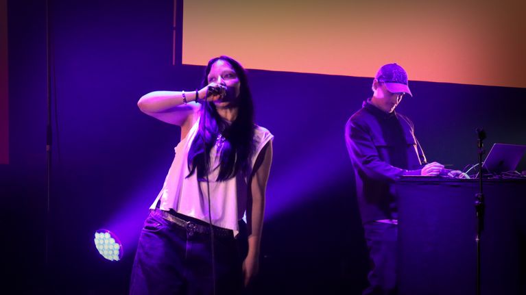Kuvassa esiintyy kaksi muusikkoa lavalla. Vasemmalla henkilö laulaa mikrofoniin ja oikealla henkilö käyttää DJ-laitteistoa.