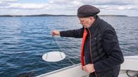 Jari Hänninen seisoo veneessä ja roikottaa veneen laidan yli narun päässä olevaa valkoista pyöreää levyä. Hänen takana näkyy meren pinta.