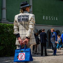 Protestoija seisoo Venäjän paviljongin edessä, hänen takissaan lukee No Putin, no war. Taustalla seisoo ihmisiä