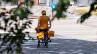 person wearing an orange helmet and shirt on a bike carrying two large mail bags on a paved road.