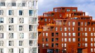 Två moderna flerbostadshus, ett vitt med perforerad fasad och ett rött med en mer slät yta, står sida vid sida under en molnig himmel. Snö täcker marken och spårvagnsspår löper framför byggnaderna.