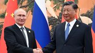 Vladimir Putin ja Xi Jinping kättelevät. Heillä on mustat puvut, Putinilla mustavalkoraitainen kravatti ja Xillä sininen, molempien kauluksessa on kullanvärinen merkki. Taustalla näkyvät Kiinan ja Venäjän liput ja seinämaalaus, jossa on kurkia.