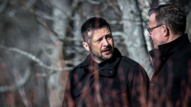 Volodymyr Zelenskyy and Petteri Orpo speaking to each other outdoors and wearing winter coats, with leafless trees in the background.