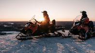 Kaksi moottorikelkkailijaa ovat pysähtyneet tunturin laelle ilta-auringon säteiden vielä osuessa heihin.