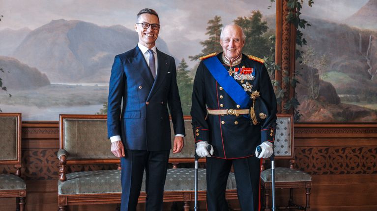 Alexander Stubb seisoo kuningas Haraldin puvussa. Harald seisoo kainalosauvojen avulla.