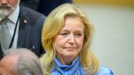 En kvinna med långt blont hår och blå blus står inomhus bland andra personer i kostym. 