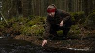 Nainen kumartuu kokeilemaan jokiveden lämpötilaa kädellään