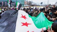 A large crowd of people holding a huge black, white, red and green flag and several smaller ones in a snowy cityscape.