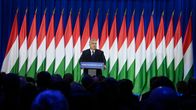 Viktor Orbán står vid ett talarpodium framför en lång rad ungerska flaggor. I förgrunden skymtar huvuden i publiken.