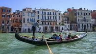 En gondol rör sig längs en kanal i Venedig. Ombord sitter tre kvinnor och en man. Gondolen ros av en stående man klädd i randig skjorta.