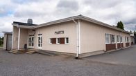 En låg byggnad med skylten "PILOTTI" på fasaden, omgiven av grusplan och några träd i bakgrunden.