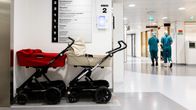Kahdet vauvanraattaat hissien luona ja taustalla kävelee kaksi henkilöä puketuneena sairaalan vihreisiin asuihin.