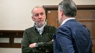 vå personer samtalar i ett rum med träpanel och klädhängare på väggen.