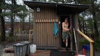 Young man only wearing green shorts walking through the front door of a small wooden sauna building, with trees and other buildings in the background.