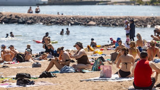 A sandy beach on a bright summer day, filled with people in bathing suits. Some are swimming in the water.