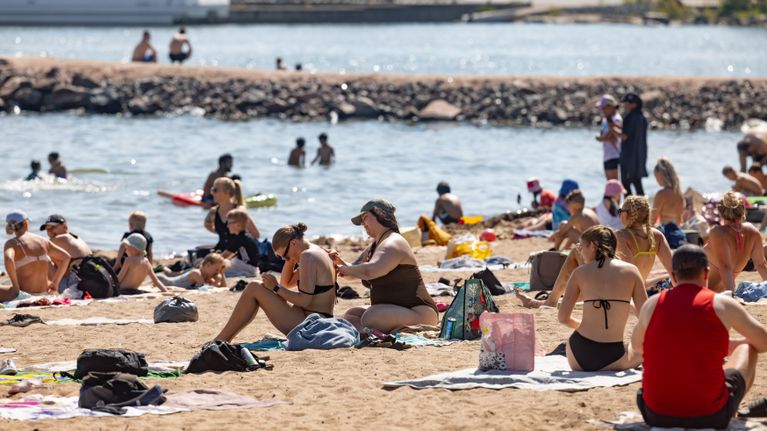 A sandy beach on a bright summer day, filled with people in bathing suits. Some are swimming in the water.