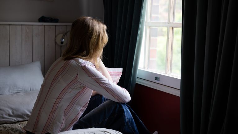 En person sitter på sängen och stirrar ut genom fönstret.