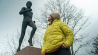 En kvinna klädd i en gul jacka står vid en skulptur och betraktar den. Skulpturen föreställer en löpare i rörelse. 