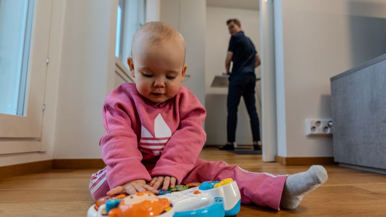 Pieni lapsi leikkii lelulla ja hänen isänsä katselee taustalla.