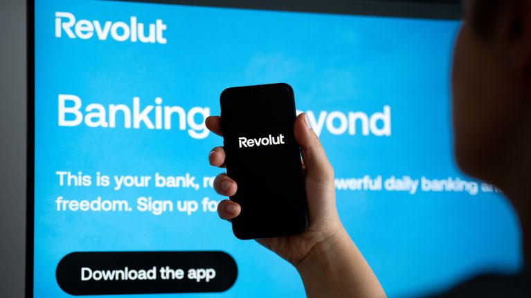 Henkilö pitää kädessään puhelinta, jonka näytössä lukee "Revolut"; taustalla tietokoneen ruudulla Revolut-mainos ja tekstiä.