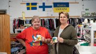 Två kvinnor står bredvid varandra framför den finländska, svenska och ukrainska flaggan, och håller varsin mugg med ukrainska symboler.