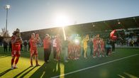 Fotbollsspelare i röda matchkläder står på en plan och applåderar inför publik på läktaren.