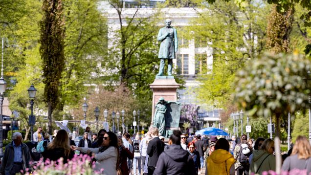 Many people walking in a leafy city park alongside two statues.