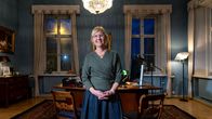 En person står i ett elegant, klassiskt inrett kontor med mörkblå väggar, antika möbler, en kristallkrona och stora fönster med gardiner.