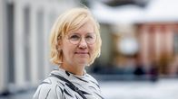 Gunilla Widén, en person med axellångt ljust hår, glasögon och en grå-vit-svart blus tittar in i kameran. Hon står utomhus, men man ser inte bakgrunden tydligt.