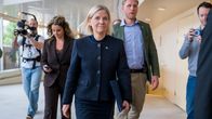 En blond kvinna i mörkblå kavaj och kjol går med en mapp i handen inomhus. 