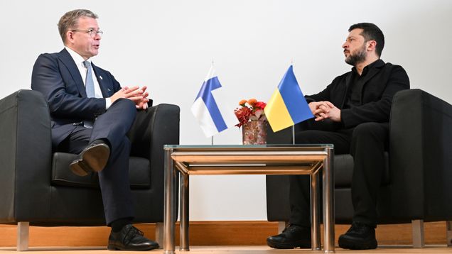 Kaksi miestä istuu tuoleilla pöydän ääressä, pöydällä Suomen ja Ukrainan liput sekä kukka-asetelma.
