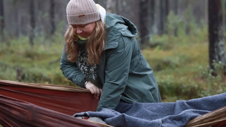 Nainen peittelee henkilöä riippumattoon metsämaisemassa.