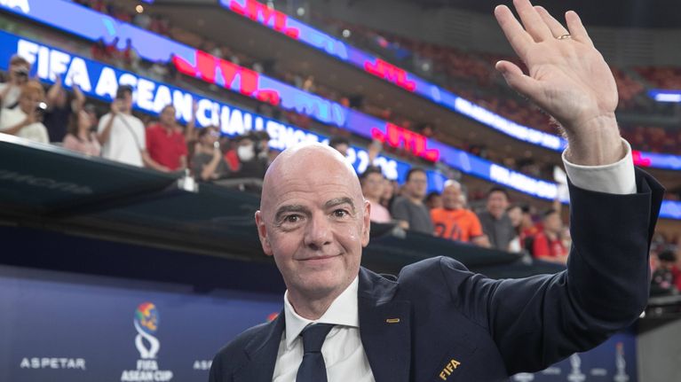 Mies puku päällään vilkuttaa kädellään katsomoon, taustalla FIFA-merkitty näyttö, jossa myös muuta tekstiä.