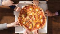 The hands of several people taking slices of pizza from a box on a table.