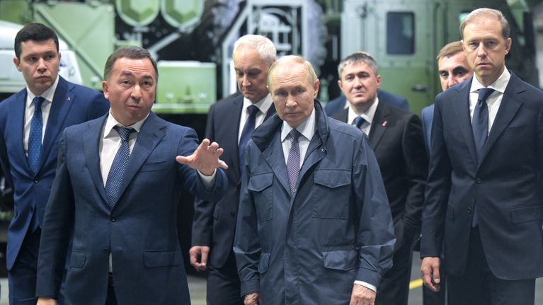 Vladimir Putin med flera kostymklädda personer i förgrunden. I bakgrunden syns att de går genom en vapenfabrik. 