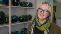 En kvinna med glasögon och färggrann skarf runt halsen står framför en hylla med färgglada glaskonstverk.
