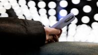 Close up photo of a person's lap, seen from the side, as the person holds a smartphone. Bright white lights are seen in the distance.