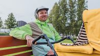 Mies istuu puistonpenkillä lippalakissa ja värikkäässä tuulitakissa. Mies työntelee keltaisia lastenrattaita ja kuuntelee hymyillen kuulokkeilla.